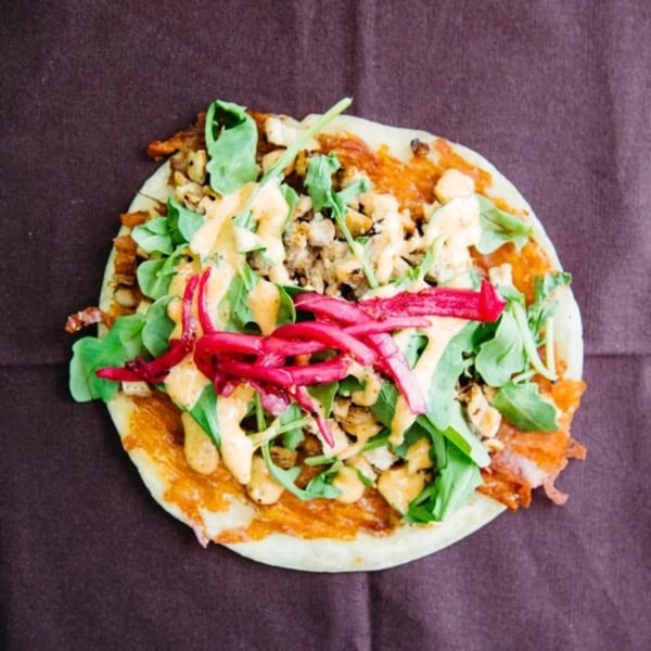 Unforked flatbread with chicken, arugula, pickled onions, and a creamy sauce on a dark surface.