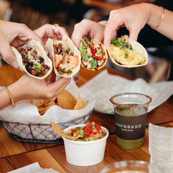 Four hands holding different tacos, with guacamole, chips, and a drink on a wooden table.