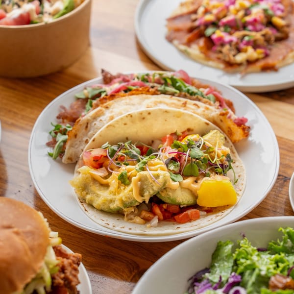A white plate of Unforked tacos with avocado, tomatoes, and microgreens, alongside a burger and salad.