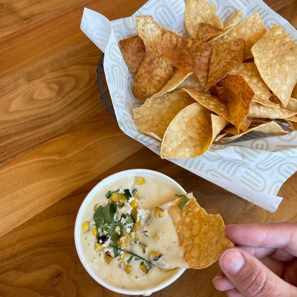 Hand dipping a tortilla chip into a bowl of Unforked elote dip with a basket of chips.