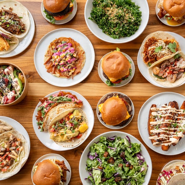 An overhead shot of a wooden table filled with various burgers, tacos, salads, and sweet potato fries.