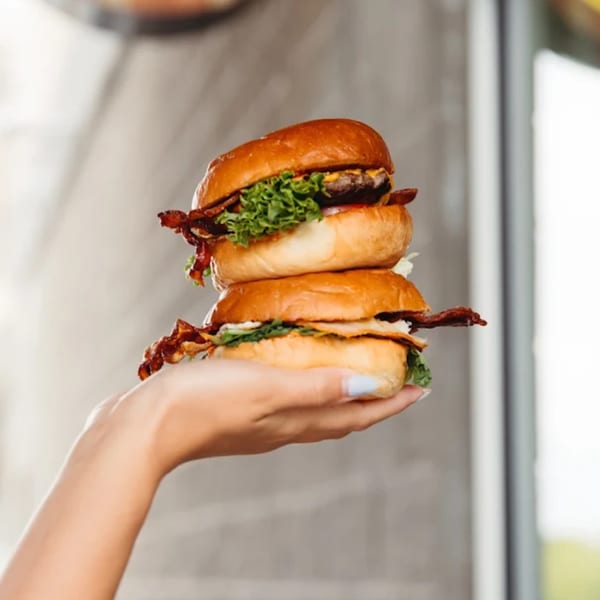 A hand holds a stack of two Unforked bacon cheeseburgers with lettuce on brioche buns.