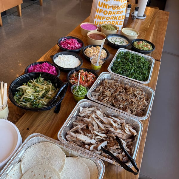 A catering spread of Unforked taco bar ingredients, including meats, tortillas, and various toppings.