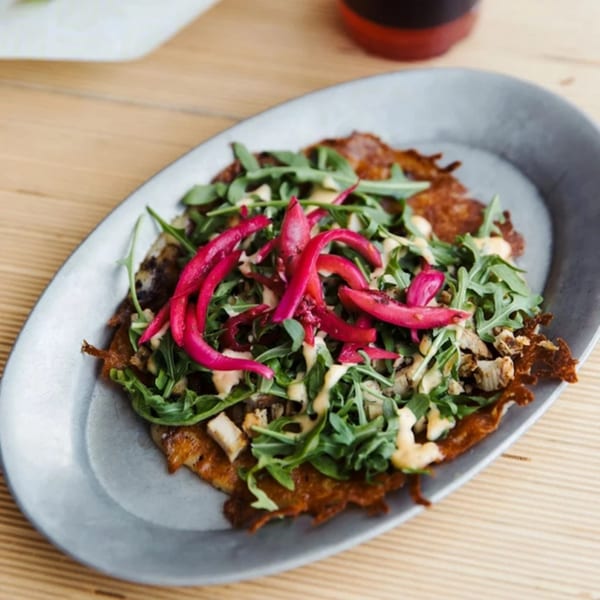 Unforked's burger patty topped with arugula, pickled red onions, and a creamy sauce on a grey plate.
