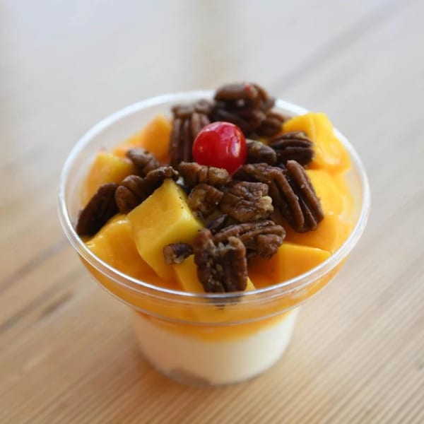 Unforked's mango dessert with pecans and a cherry in a clear cup on a wooden table.