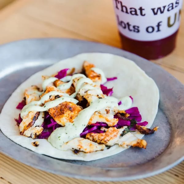 Unforked chicken taco with purple cabbage and white sauce on a metal plate, with a drink in the background.