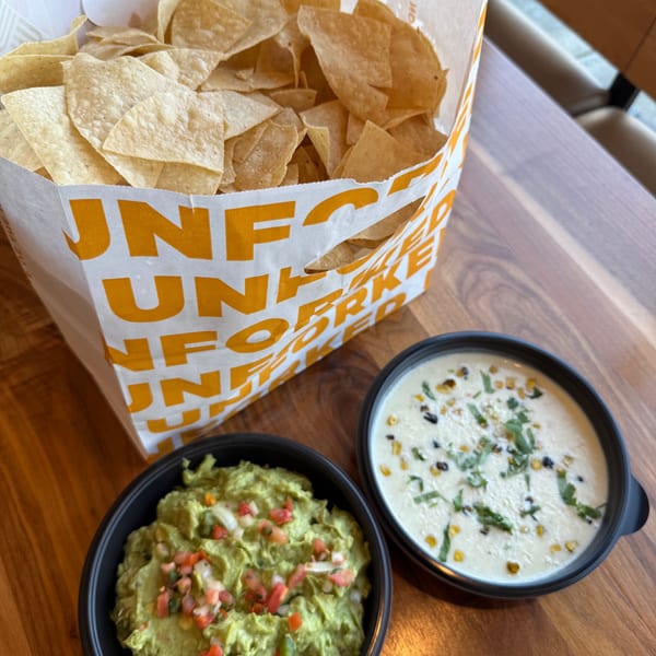 Unforked tortilla chips in a bag with guacamole and queso dips on a wooden table.
