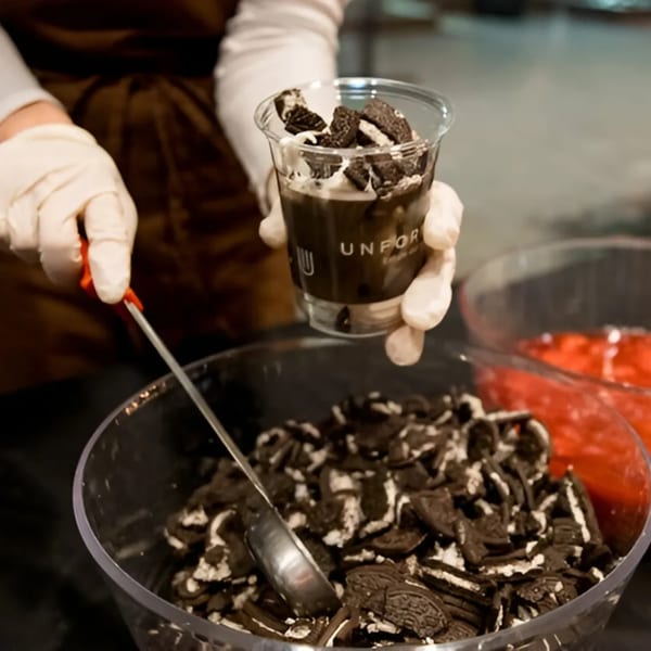 A gloved hand holds an Unforked dessert cup filled with chocolate and Oreo crumbles, next to a bowl of more Oreos.