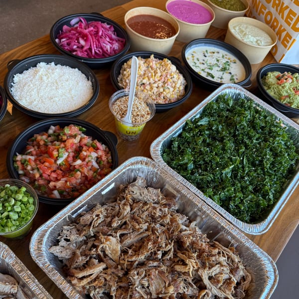 A catering spread of Unforked burger toppings, including pulled pork, kale, rice, and various sauces.