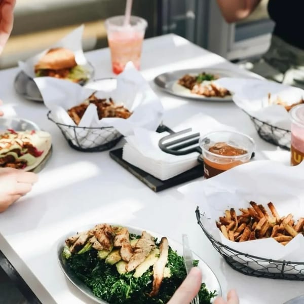 A table filled with Unforked burgers, fries, salads, and drinks, with people's hands reaching for food.